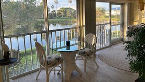 A balcony with a glass table and two chairs, overlooking a body of water and greenery.