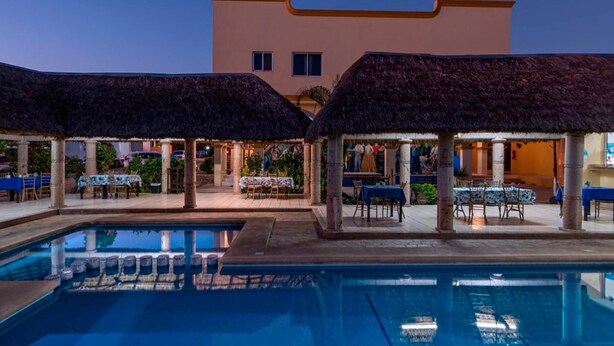 A poolside area with thatched umbrellas, tables, and chairs, and a building in the background. - Hotel Yauco (Rosario)
