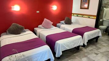 Habitación de hotel con tres camas, cada una con una colcha morada y un almohadón mullido. Hay luces empotradas en la pared roja y una obra de arte enmarcada sobre la puerta.
