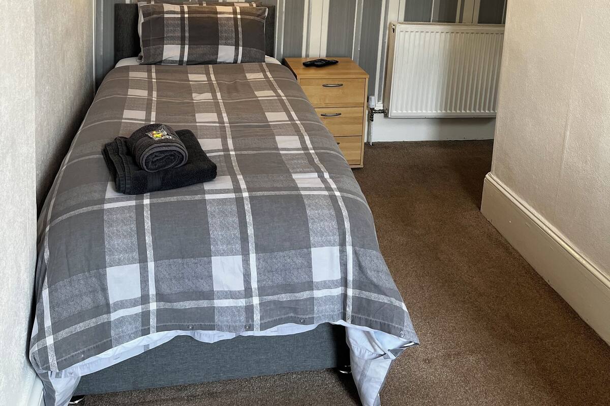 A neatly made bed with a plaid bedspread, a bedside lamp, and a small wooden nightstand with a remote control.
