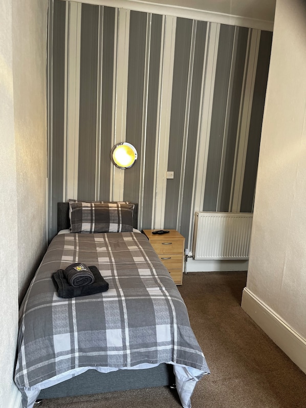 Un lit bien fait, recouvert d’une couverture à carreaux, une lampe de chevet et une petite table de nuit en bois avec une télécommande.