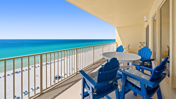 A balcony with a view of the beach, a round table, and blue chairs.