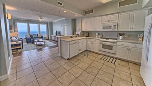 A spacious kitchen with white cabinets, a microwave, and a stove. There is a dining area with a table and chairs, and a living room with a sofa and a view of the ocean.