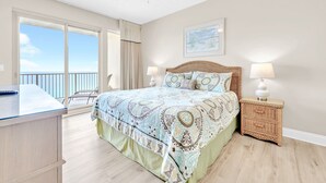 A bedroom with a large bed, a ceiling fan, a nightstand, a chair, and a view of the ocean.