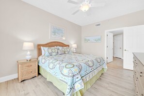 Un dormitorio con una cama, dos mesitas de noche, un ventilador de techo y una puerta que da a otra habitación.