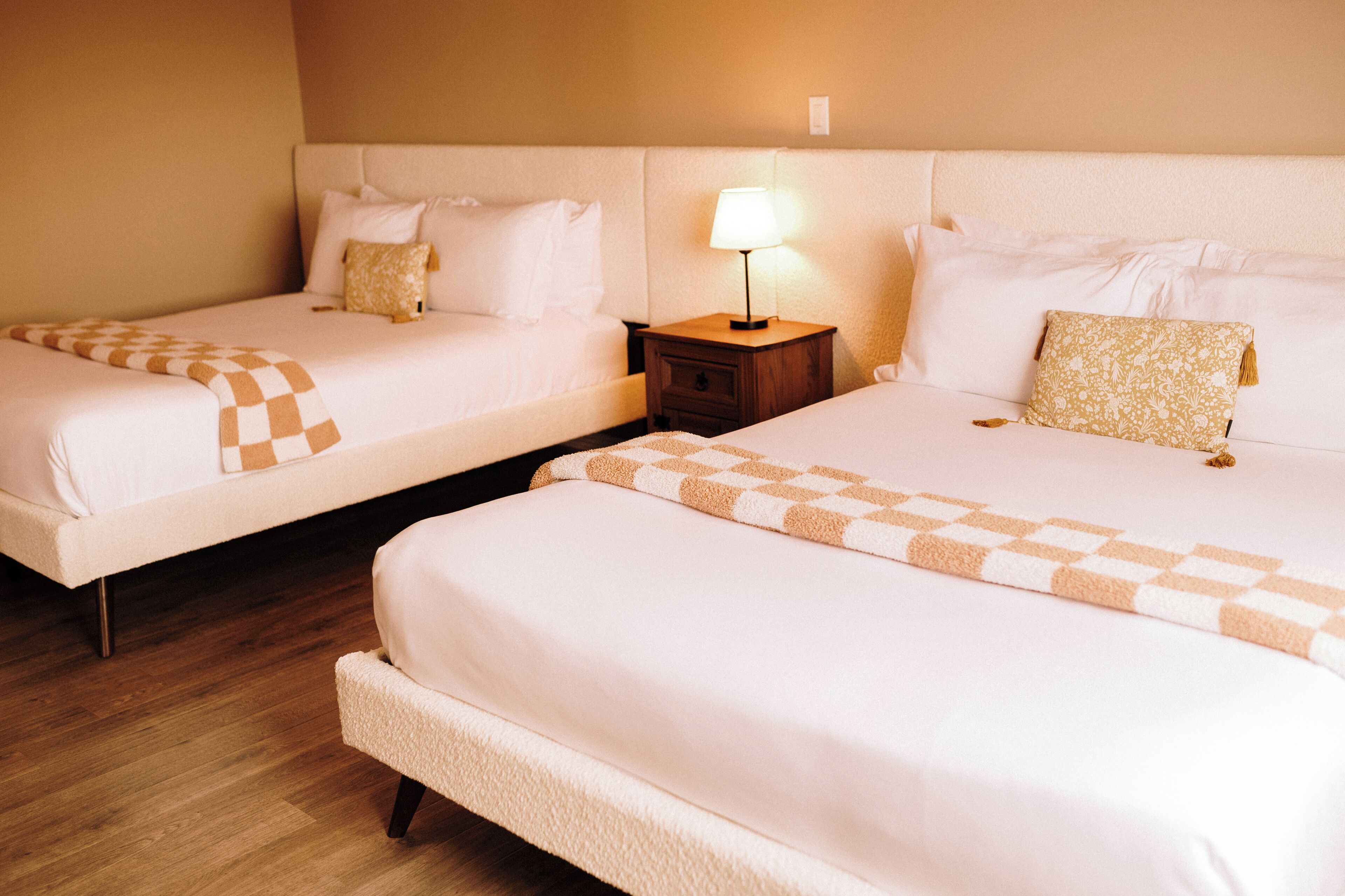 Two single beds with checkered blankets, a nightstand with a lamp, and a wooden floor.