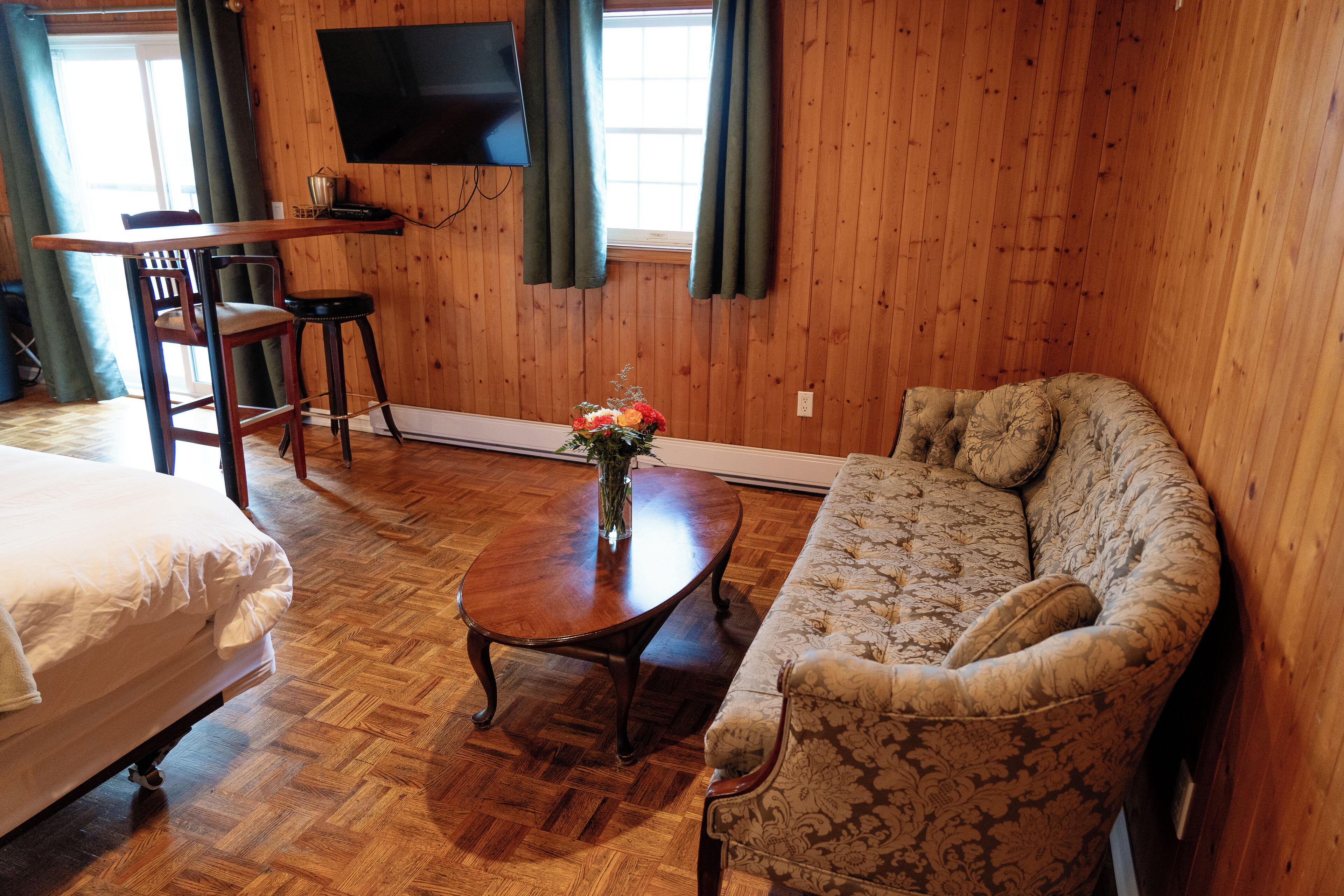 A wooden-paneled room with a bed, a sofa, a dining table with chairs, a TV mounted on the wall, and a window with curtains.