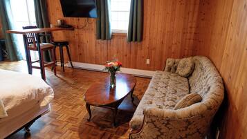 A wooden-paneled room with a bed, a sofa, a dining table with chairs, a TV mounted on the wall, and a window with curtains.