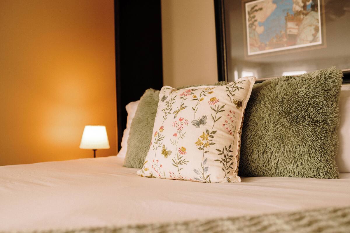 Ein ordentlich gemachtes Bett mit blumigen und grünen Kissen, einer Lampe daneben und einem gerahmten Bild an der Wand.