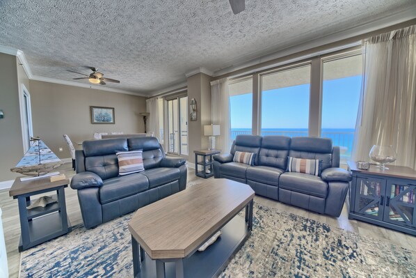 A spacious living room with a large window offering a view of the ocean, a ceiling fan, and a dark wood coffee table.