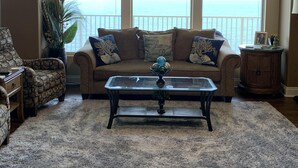 A living room with a sofa, armchair, and coffee table, large windows with a view of the ocean, and a ceiling fan.