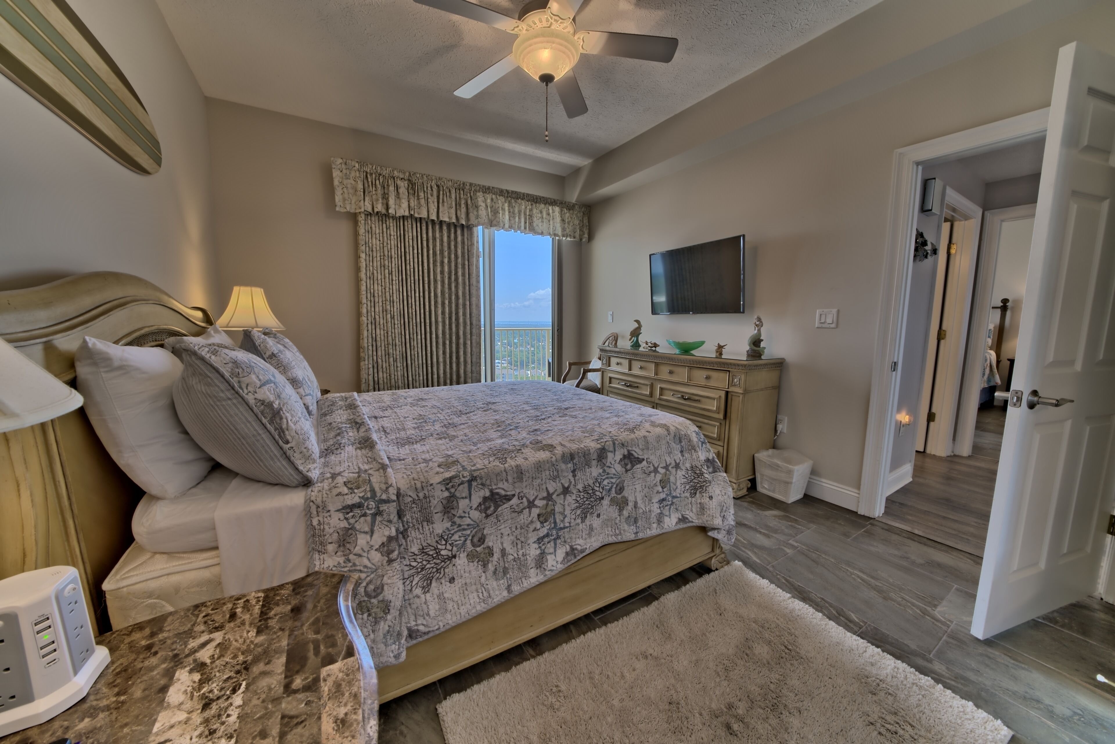 A bedroom with a bed, a TV, a ceiling fan, a nightstand, and a view of the outdoors.