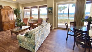 A spacious living room with a large sofa, a dining table with chairs, a wooden cabinet, and a view of the ocean through large windows. - Gulf-Front Escape | 1st Floor + 2BR (Panama City Beach)