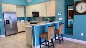 A kitchen with teal walls, white cabinets, a stainless steel refrigerator, a microwave, a sink, and two leopard print bar stools. - Gulf-Front Escape | 1st Floor + 2BR (Panama City Beach)