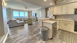 A spacious living area with a gray sectional sofa, a gray armchair, and a dining table. The kitchen features white cabinets, a stainless steel sink, and a built-in oven. - Gulf Crest 2304 | Your Gulf-Front Home Away From Home! (Panama City Beach)