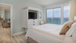 A bedroom with a bed, a TV, a dresser, a chair, and a view of the ocean.