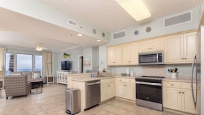 A modern kitchen with light-colored cabinets, a stainless steel refrigerator, a microwave, and an oven. There is a dining area with a sofa and a TV.