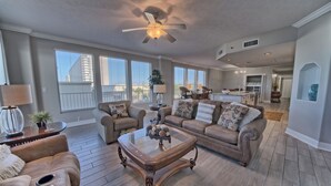 A spacious living room with a large window, a ceiling fan, and a kitchen in the background. - 3BR Gulf-Front Retreat | Steps to the Sand (Panama City Beach)