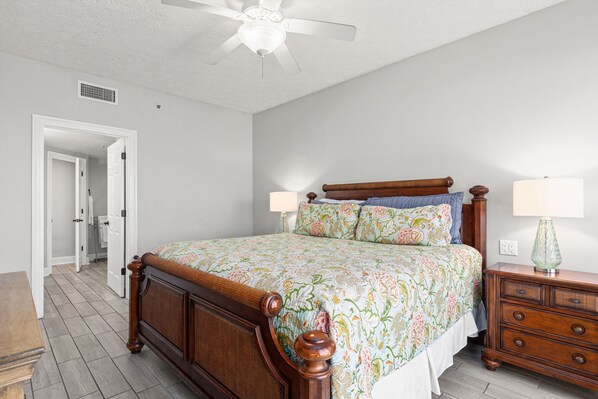 A bedroom with a wooden bed, two bedside tables with lamps, a ceiling fan, and a door leading to another room.