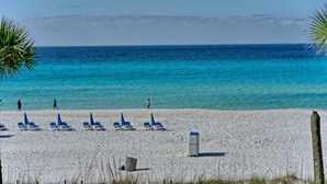 A beach with white sand, clear blue water, and a few people walking. There are blue umbrellas and lounge chairs arranged on the sand. - 3BR Gulf-Front Retreat | Steps to the Sand (Panama City Beach)