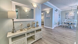 A spacious living area with a wooden floor, a white console table with decorative items, a large mirror, and a dining area with a white table and chairs. - Gulf Crest 301 – 3BR Haven with 360° Views of PCB! (Panama City Beach)