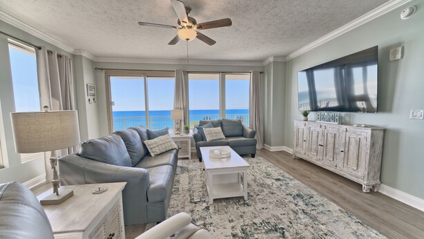 A spacious living room with a large window overlooking the ocean, a ceiling fan, and a flat-screen TV mounted on the wall. - Gulf Crest 301 – 3BR Haven with 360° Views of PCB! (Panama City Beach)