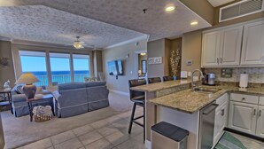 A modern kitchen with a central island, a dining area with a gray sofa, and a view of the ocean. - Gulf Crest 606: Escape to a Cozy 2-Bedroom with Gulf Views! (Panama City Beach)