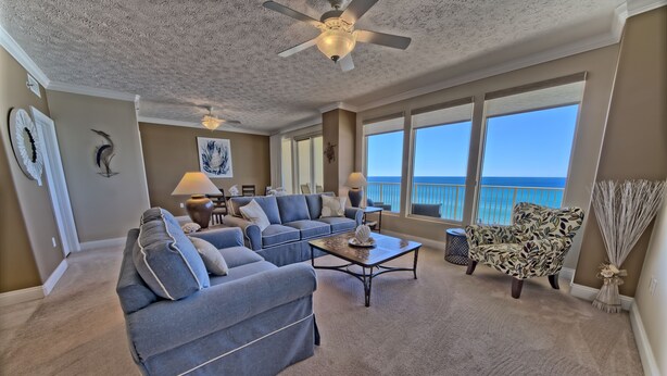 A spacious living room with a large blue sofa, a patterned armchair, and a view of the ocean through large windows. - Gulf Crest 606: Escape to a Cozy 2-Bedroom with Gulf Views! (Panama City Beach)