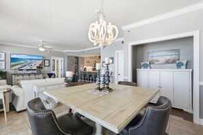 A dining area with a wooden table, black upholstered chairs, a chandelier, and a view of the beach through a large window. - Relax at Gulf Crest: 2-Bedroom Condo Bella! (Panama City Beach)