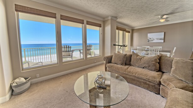 Una sala de estar con una amplia mesa de centro de vidrio, alfombra color beige y un sofá beige. Hay ventanas grandes con vista a la playa y un ventilador de techo.