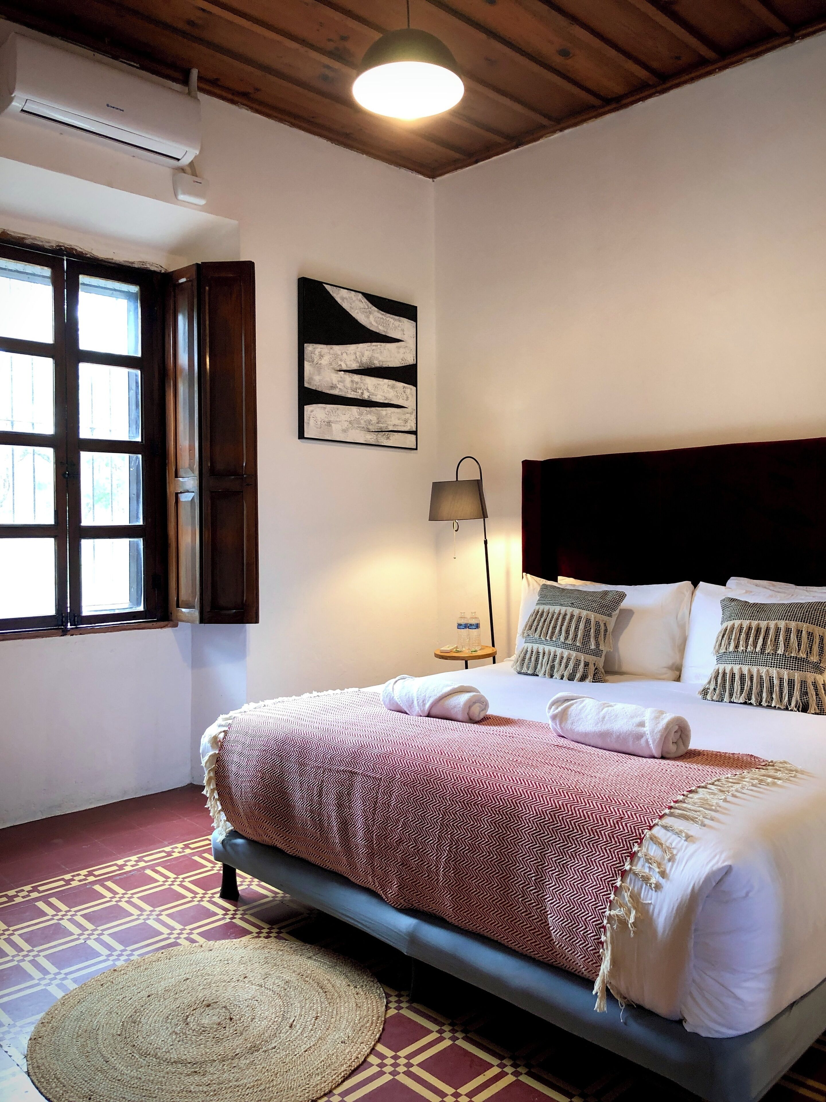 A hotel room with a bed, a wall-mounted air conditioner, a window with wooden shutters, a round rug, and a wall art piece.