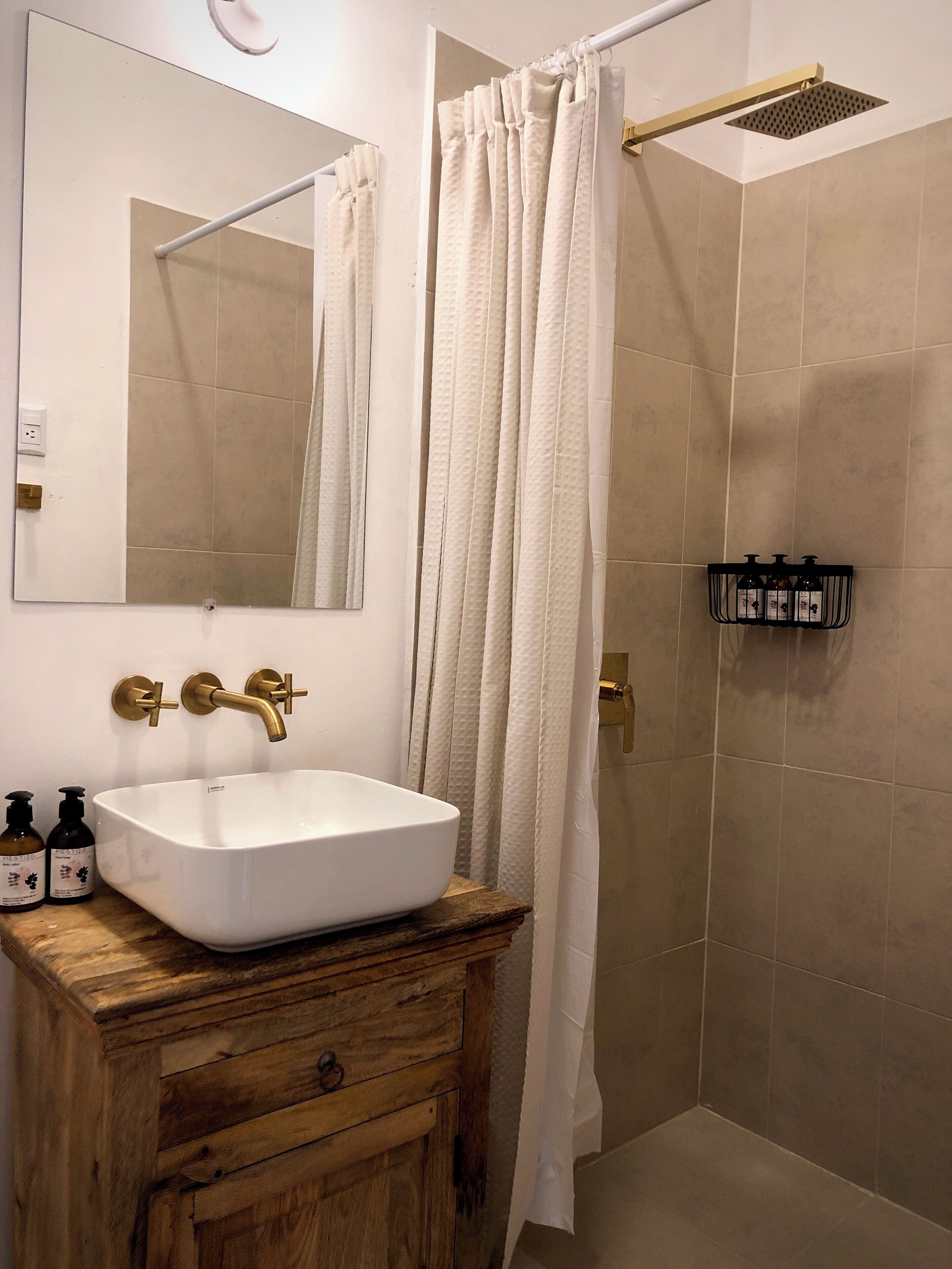 A bathroom with a white sink, a wooden vanity, a mirror, a shower with a white curtain, and a shelf with toiletries.