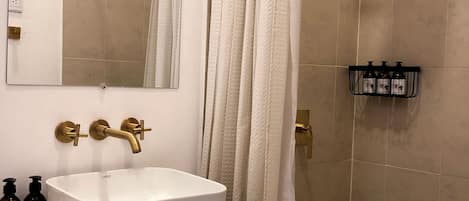 A bathroom with a white sink, a wooden vanity, a mirror, a shower with a white curtain, and a shelf with toiletries.