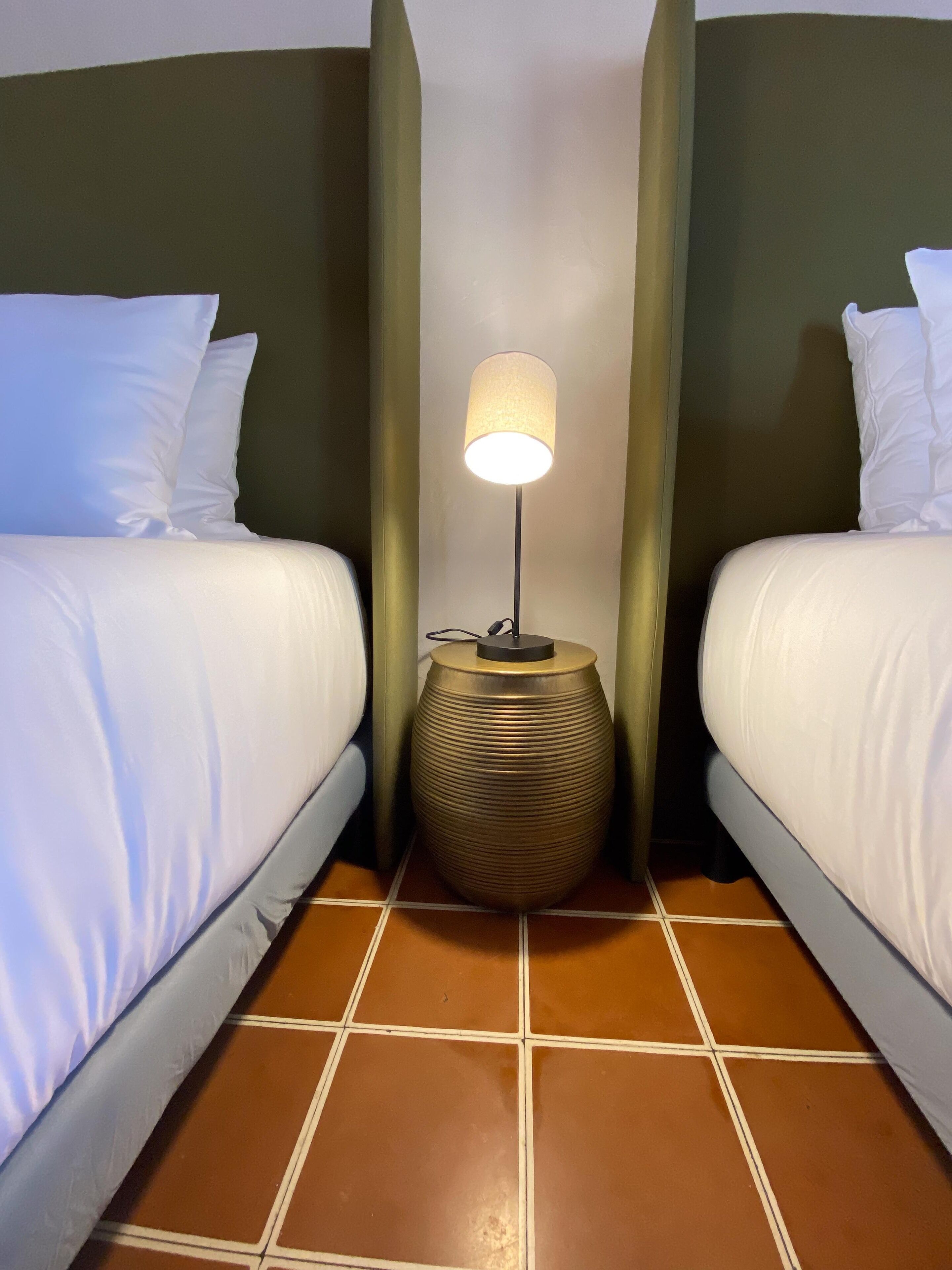 Habitación de hotel con dos camas, una mesita de noche con lámpara y piso de baldosas.
