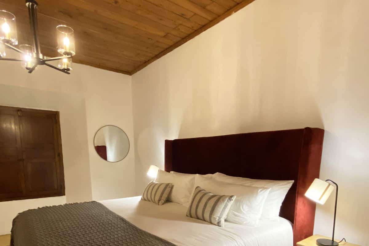 Un dormitorio con techo de madera, cama con cabecera roja, lámpara de noche, espejo y baldosas de suelo con diseño.