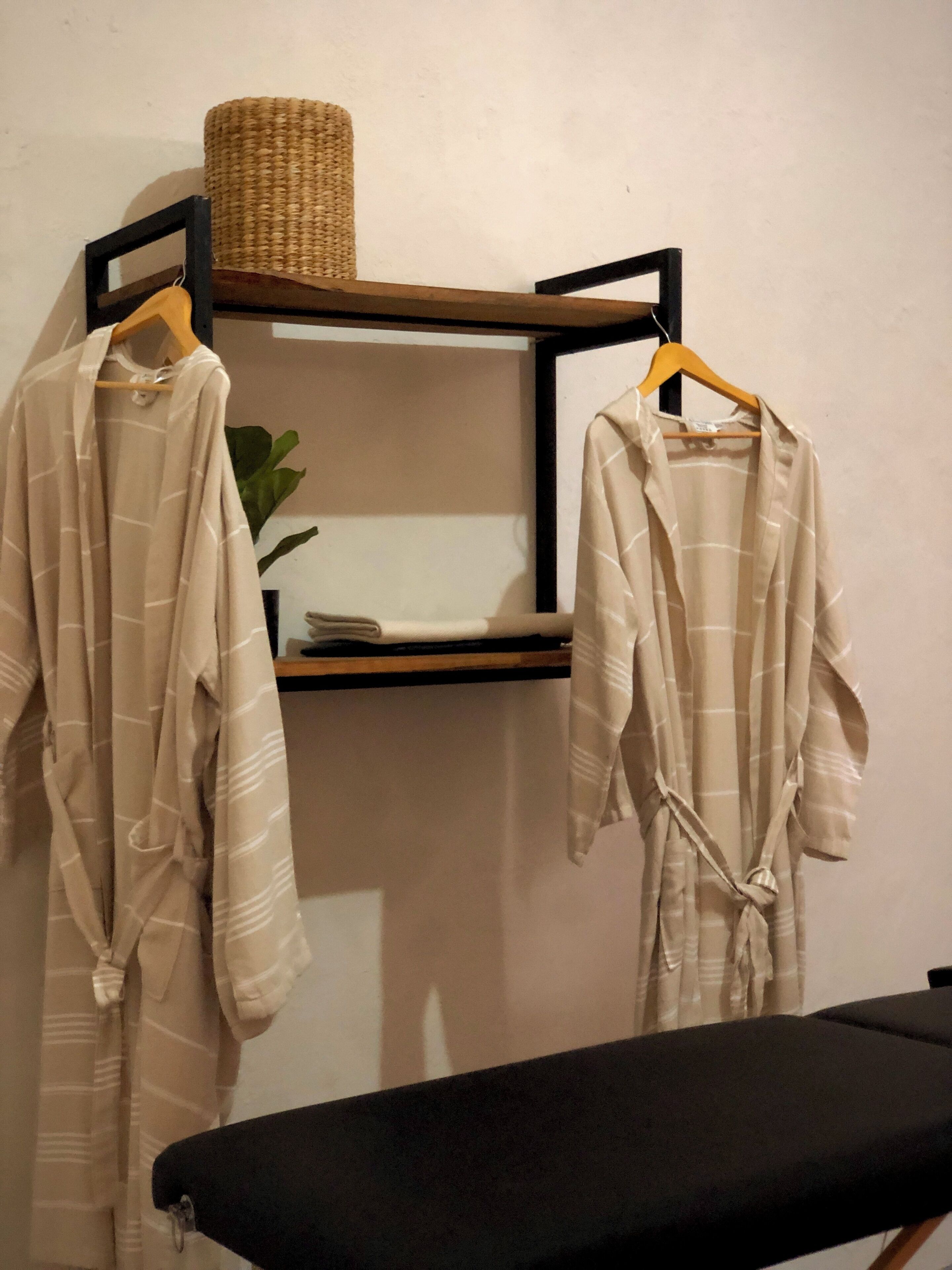 A room with a black massage table, a shelf with a woven basket, and two robes hanging on a rack.