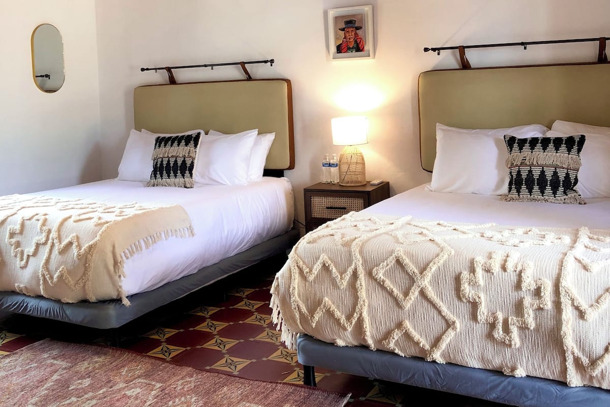 Habitación de hotel con dos camas, techo de madera, suelo estampado y una mesita de noche con lámpara.