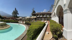 Una piscina con una fuente central, rodeada de setos cuidados y un edificio de arquitectura clásica.