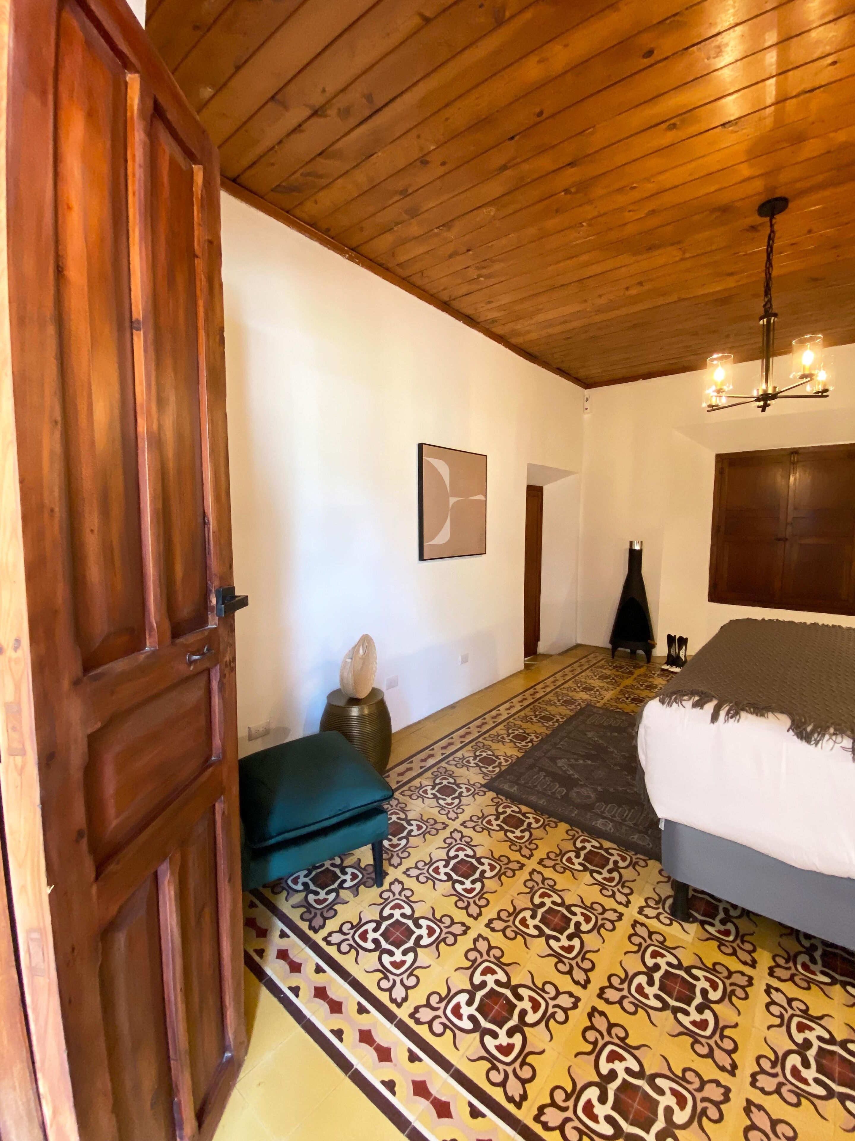 Una habitación con techo de madera, una cama con manta gris, un tapete estampado, una puerta de madera y un aplique de pared.