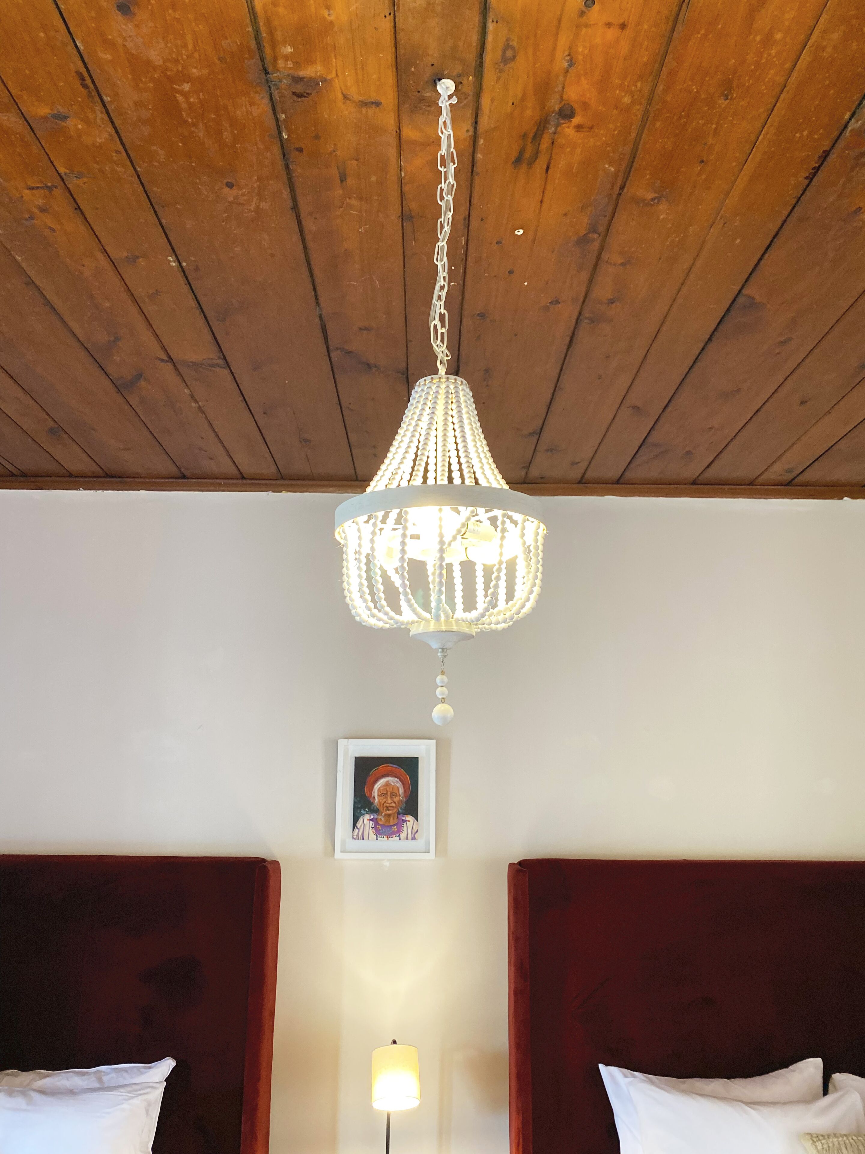 Une pièce avec des poutres apparentes en bois au plafond, un lustre blanc, un tableau encadré au mur et deux lits avec des oreillers blancs.