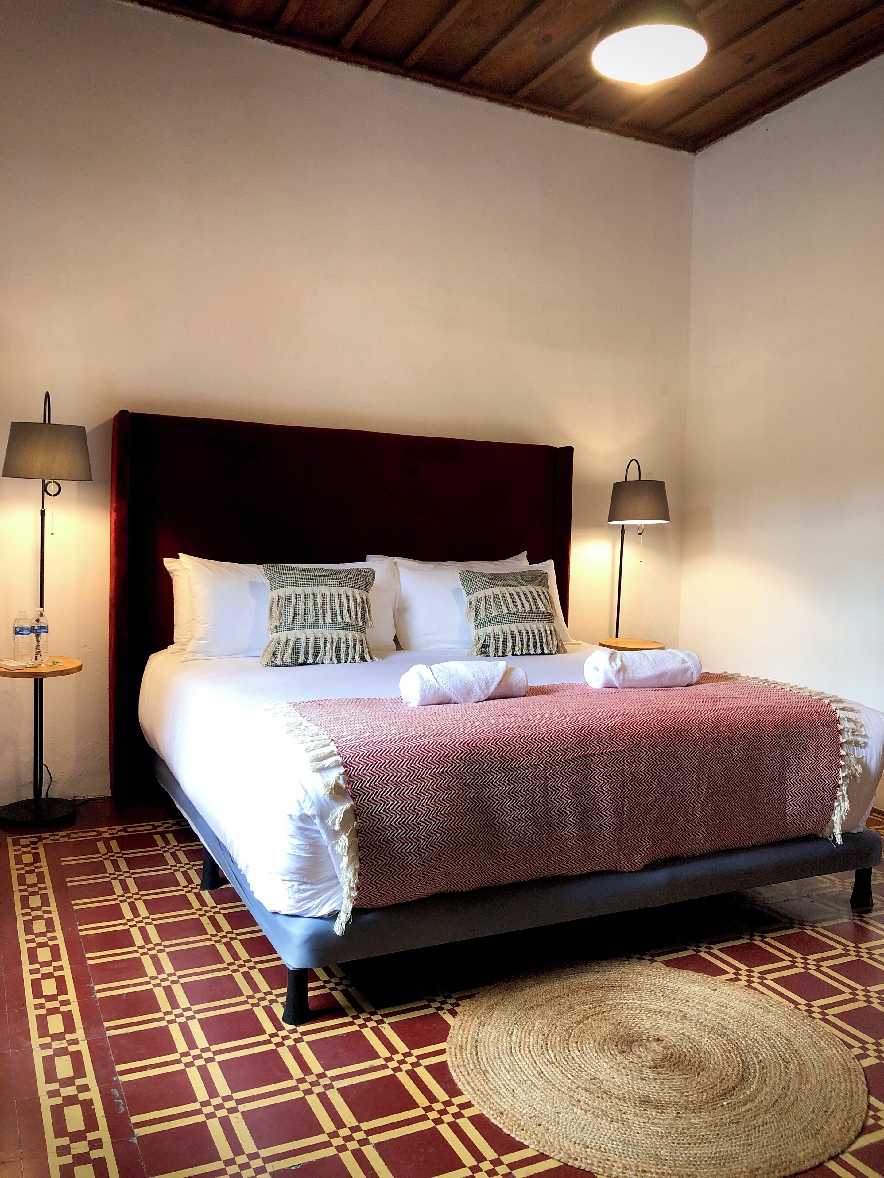 Una cama bien hecha con una colcha de cuadros rojos y blancos, dos lámparas de noche y una alfombra tejida sobre un suelo estampado.