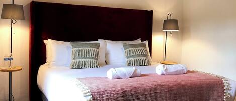 Una cama bien hecha con una colcha de cuadros rojos y blancos, dos lámparas de noche y una alfombra tejida sobre un suelo estampado.
