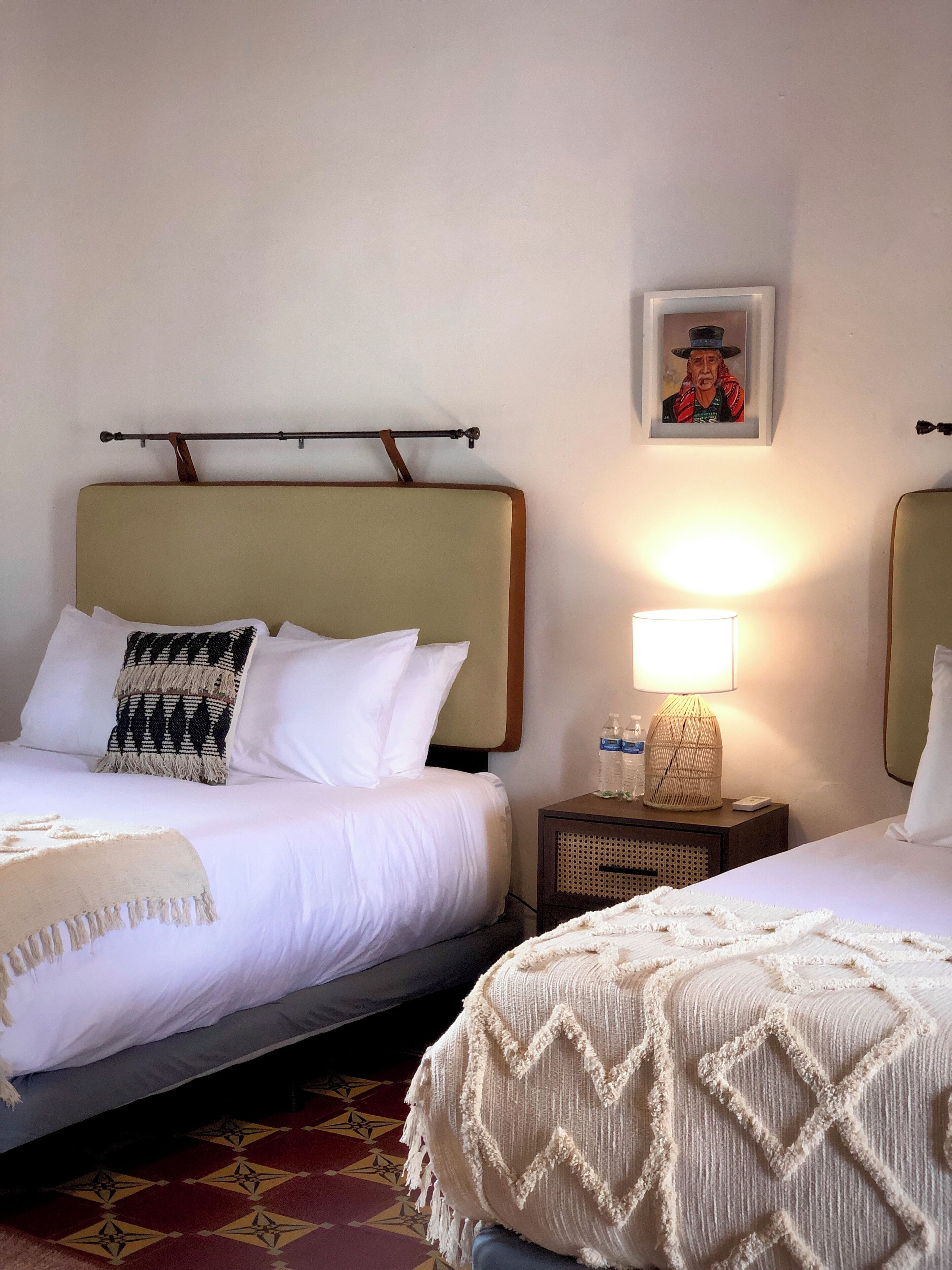 Une chambre d’hôtel avec deux lits, une table de chevet avec une lampe, un tableau encadré au mur et un tapis à motifs.