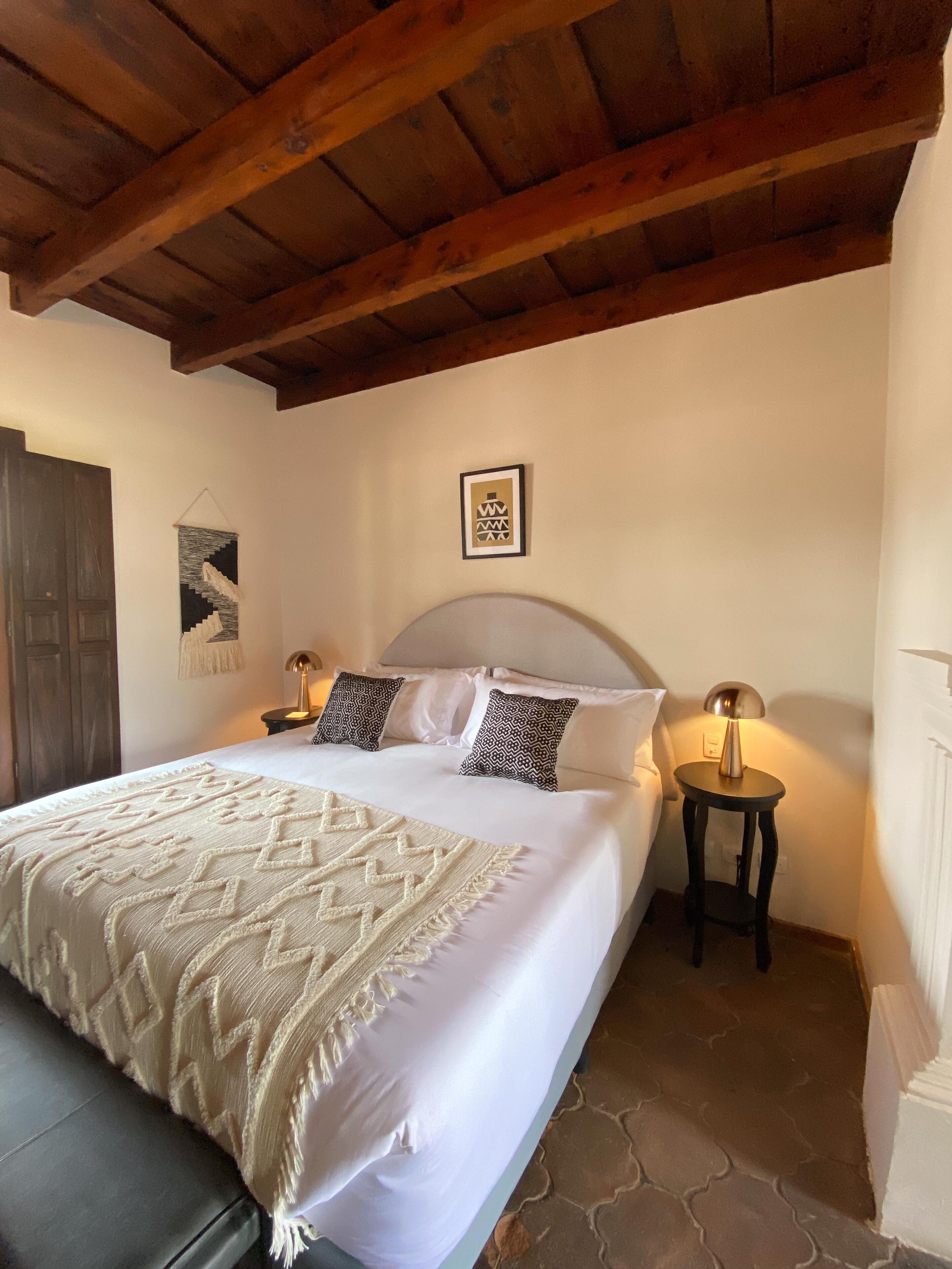 Un dormitorio con una cama grande, dos mesitas de noche con lámparas, techo de madera y un cuadro en la pared.