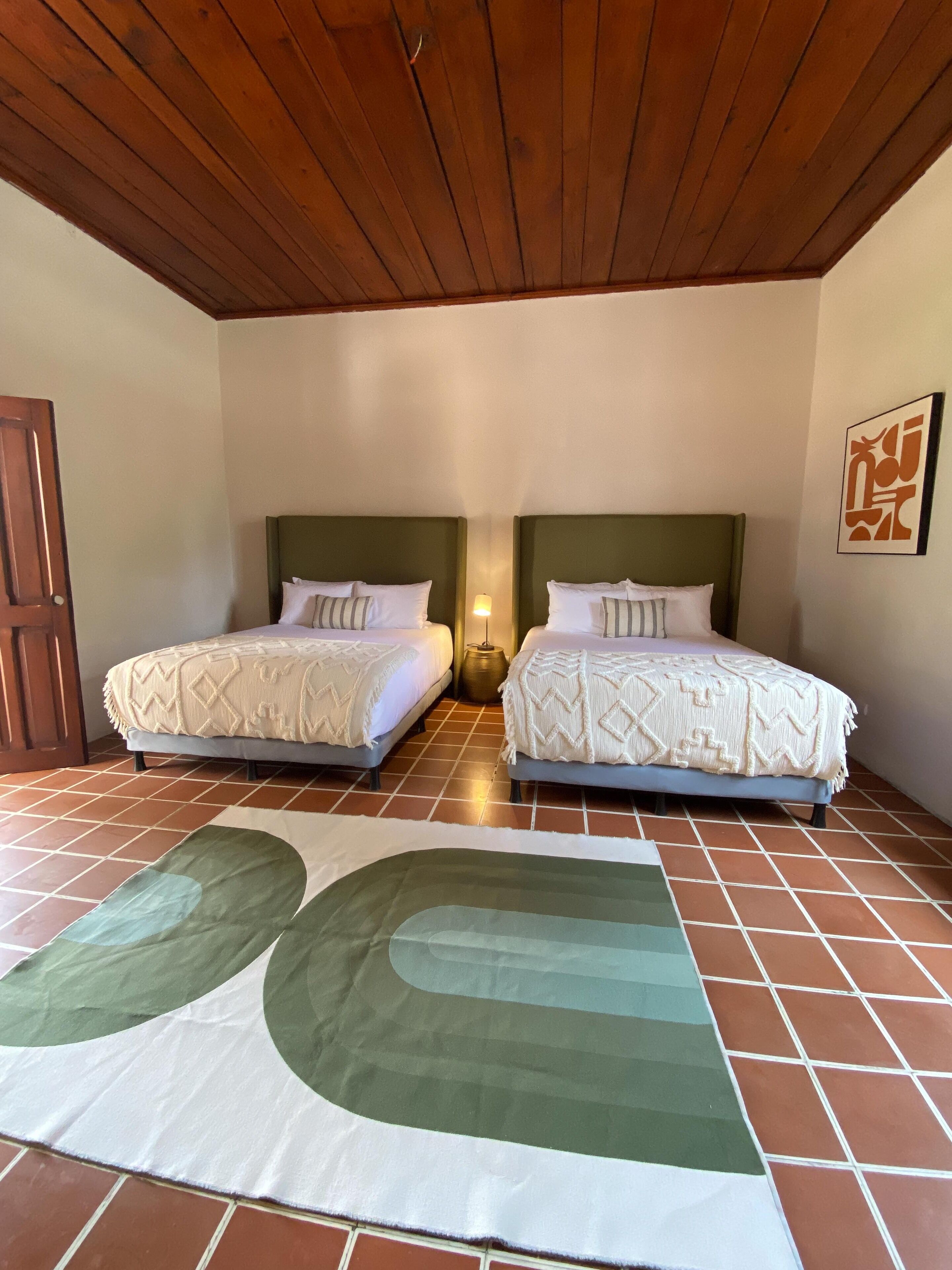 Habitación de hotel con dos camas, techo de madera, suelo de baldosas y una alfombra grande de diseño abstracto.