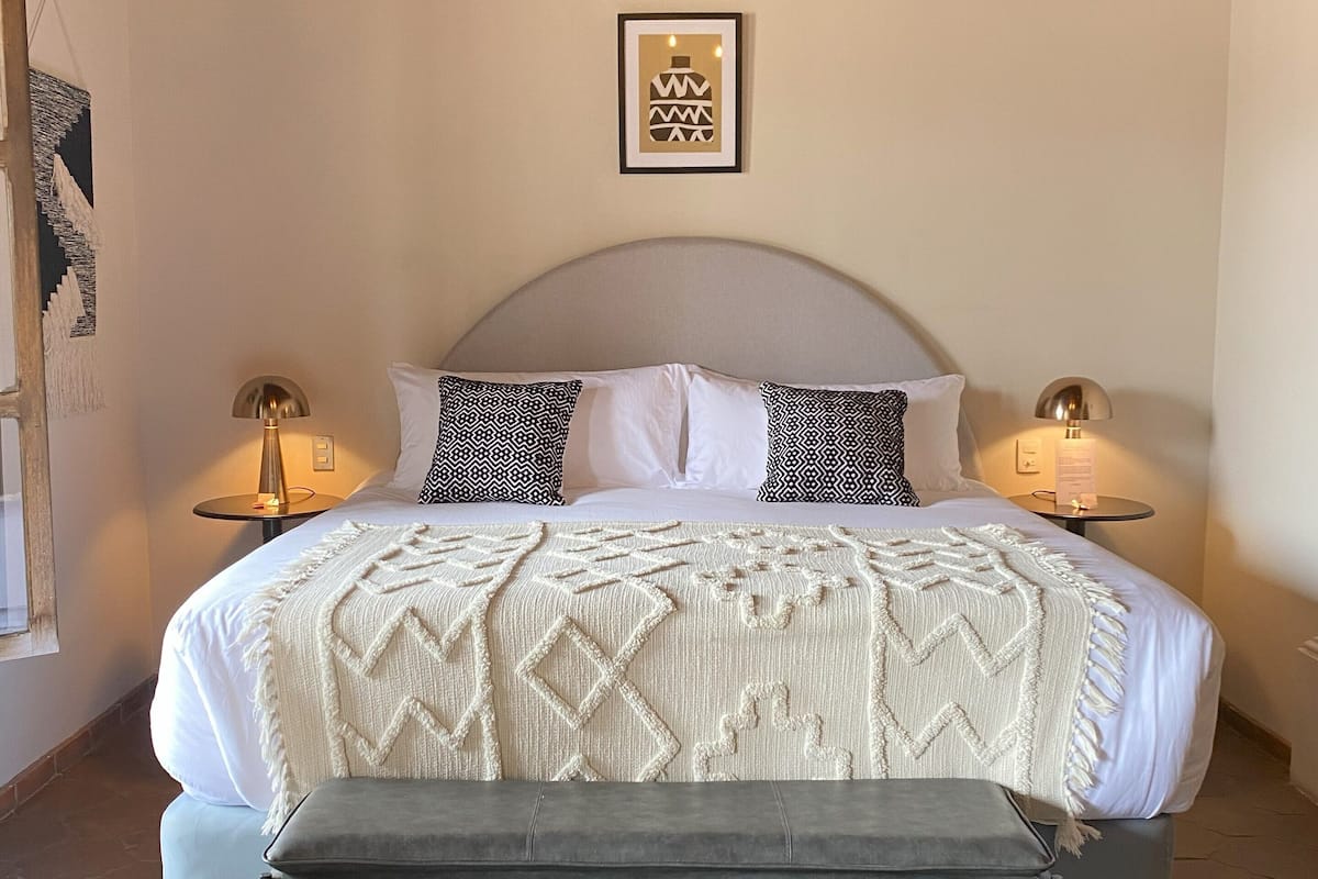 Un dormitorio ordenado con cama, lámparas de noche, un banco y una alfombra estampada.