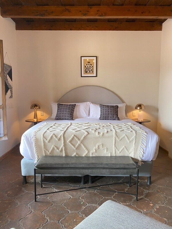 Un dormitorio ordenado con cama, lámparas de noche, un banco y una alfombra estampada.