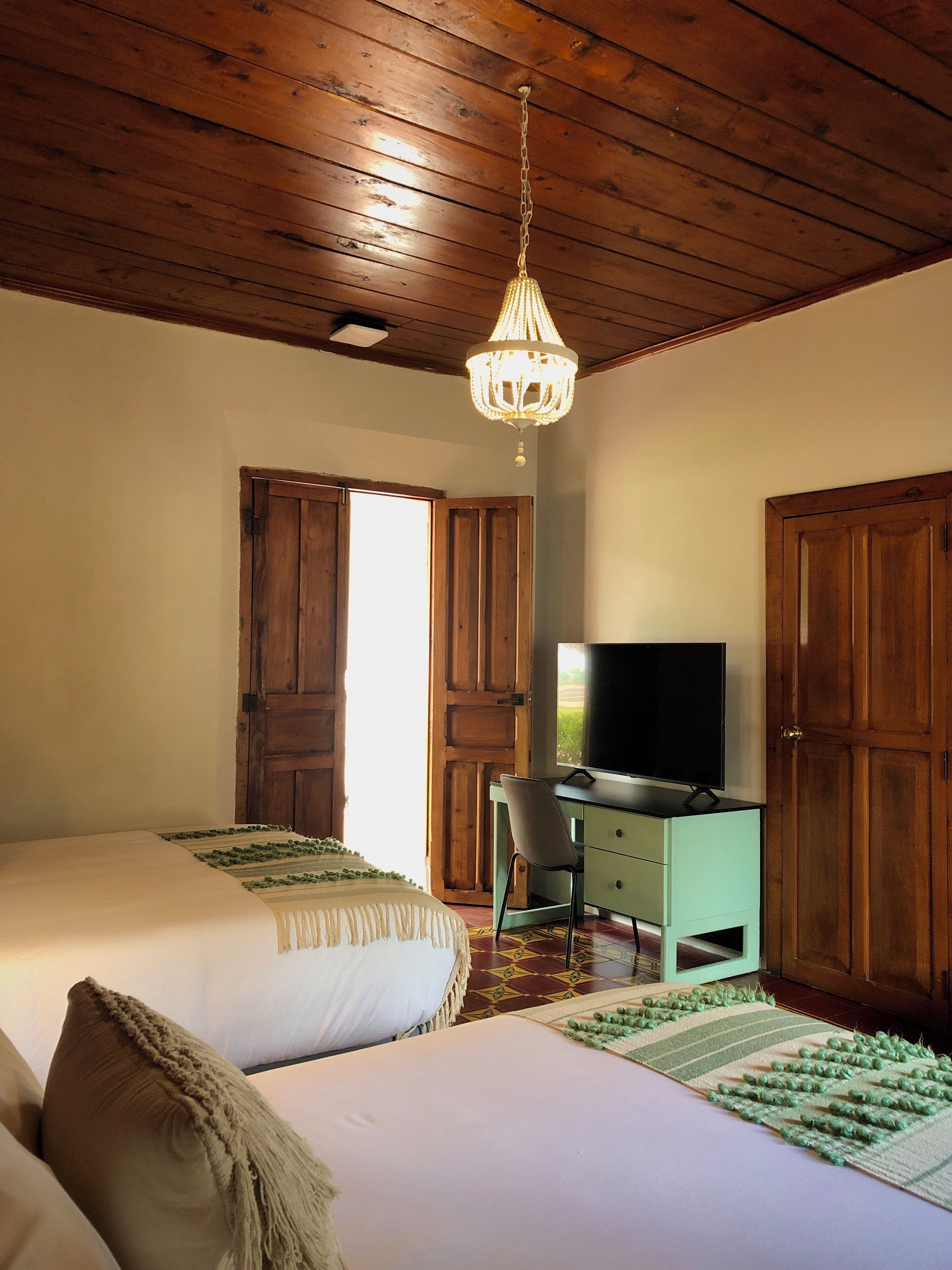 A bedroom with two beds, a desk with a TV, wooden doors, and a chandelier.
