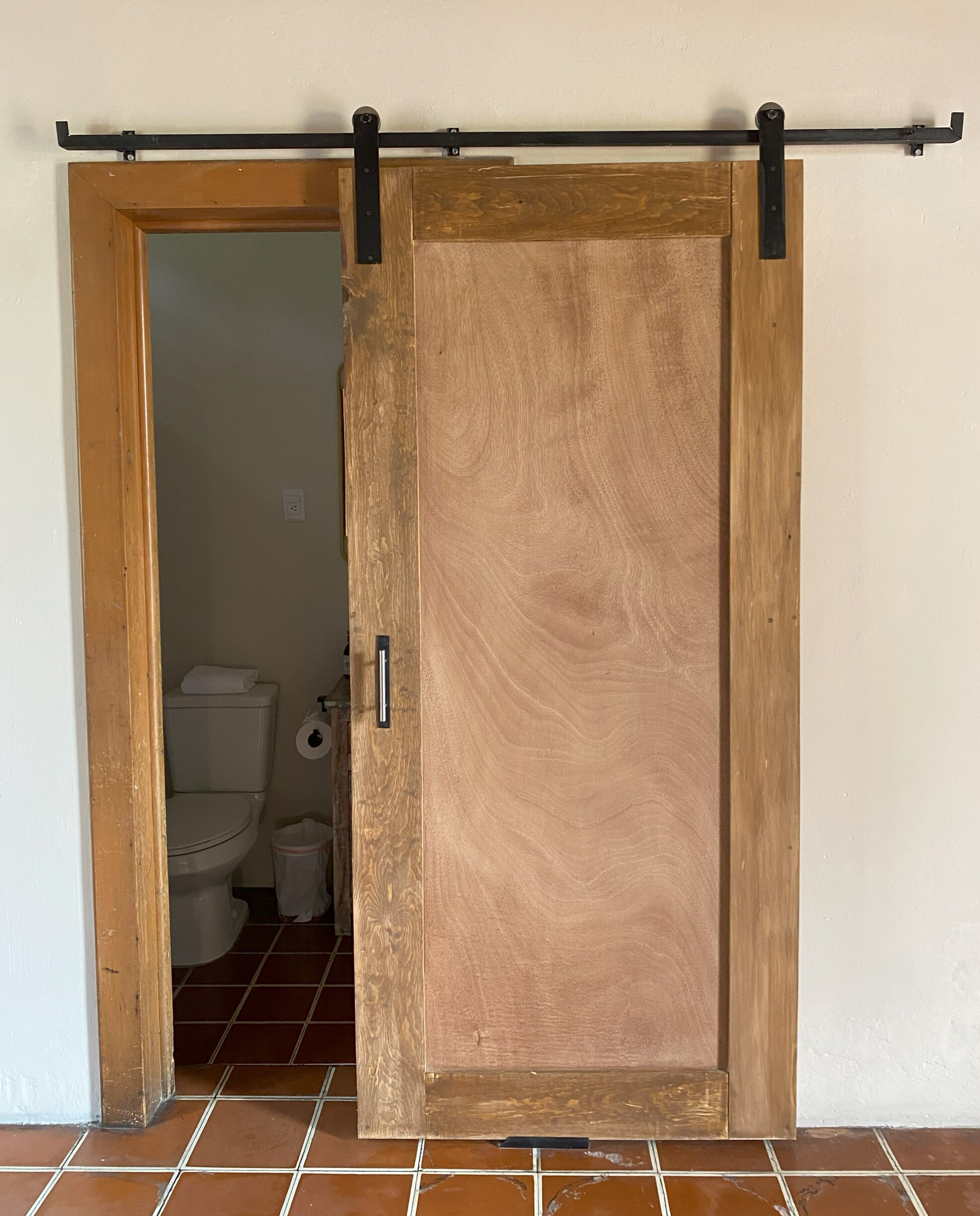 A wooden sliding door with a metal track, leading to a bathroom with a toilet and a trash bin.