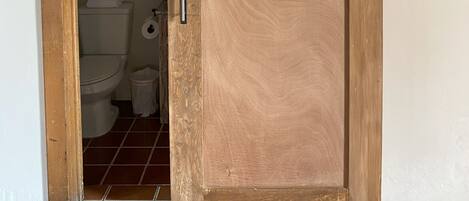 A wooden sliding door with a metal track, leading to a bathroom with a toilet and a trash bin.